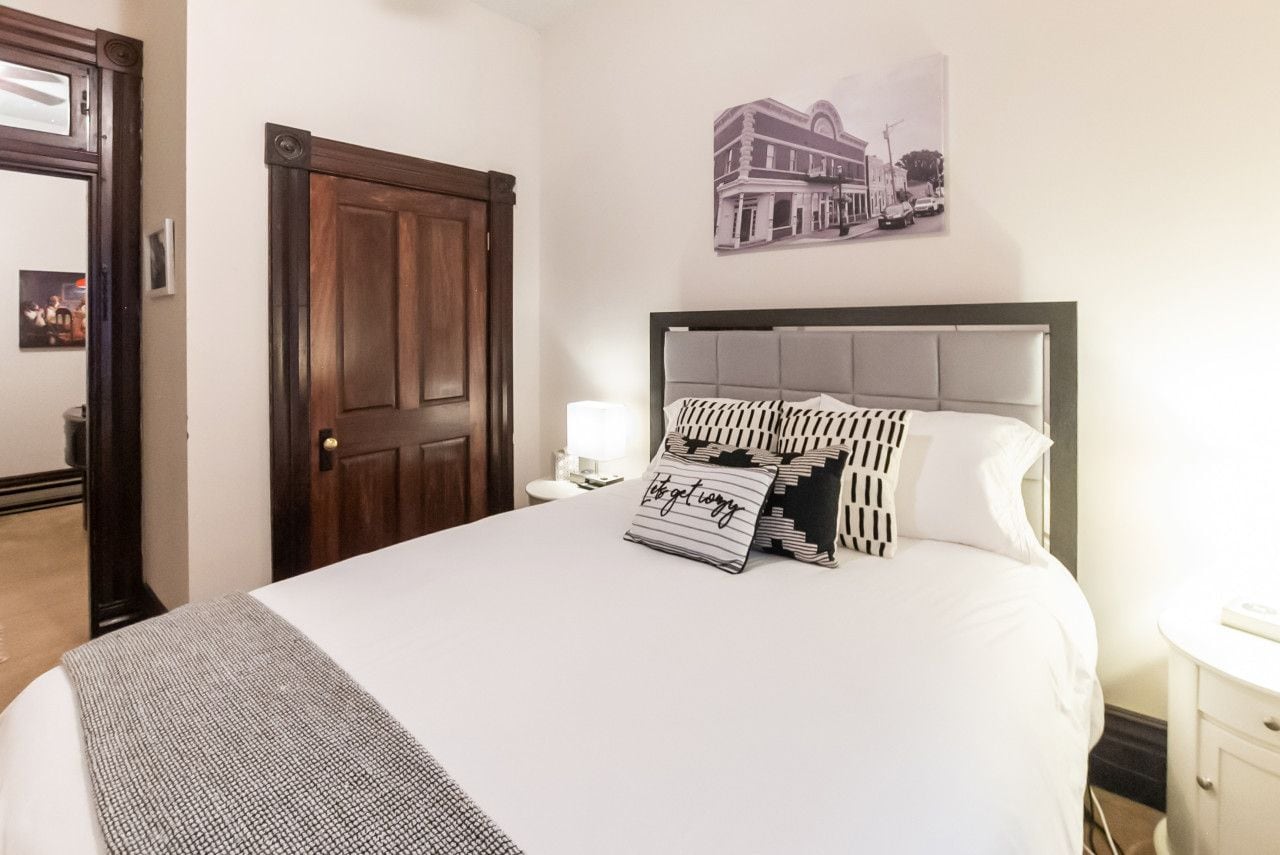 A hotel room with a large bed, a wooden door, a wall-mounted picture, and a bedside table with a lamp.
