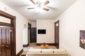 A hallway with a ceiling fan, a flat-screen TV mounted on the wall, a wooden coffee table with a plant, and a beige sofa.