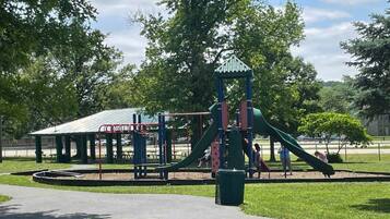 A playground with a slide and climbing structures, surrounded by grass and trees.