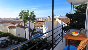 Un balcón con una mesa de madera y un cuenco de fruta, con vistas a una zona residencial con edificios blancos y un cielo azul despejado.