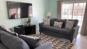 A living room with a grey sofa, a black entertainment unit, a TV, and a window with curtains. - Blue SeaShell (North Myrtle Beach)