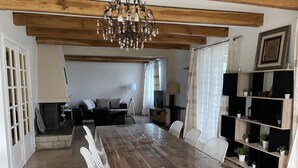 Un comedor con una mesa de madera, sillas blancas y una araña. Hay un sofá y un televisor al fondo.