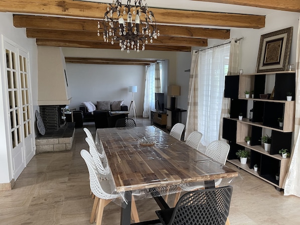 Een eetgedeelte met een houten tafel, witte stoelen en een kroonluchter. Achterin staat een bank en een televisie.