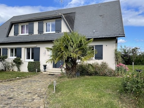 Ein zweistöckiges Haus mit einem geneigten Dach, einem steinernen Weg zum Eingang und einem gepflegten Garten mit verschiedenen Pflanzen und Blumen.