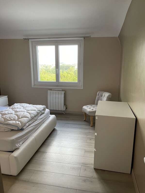 Een slaapkamer met een bed, een stoel, een radiator en een raam met uitzicht op groen.