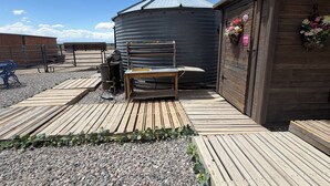 Ett rustikt utomhusområde med ett trädäck, en metallsilo och en träbyggnad dekorerad med blommor.