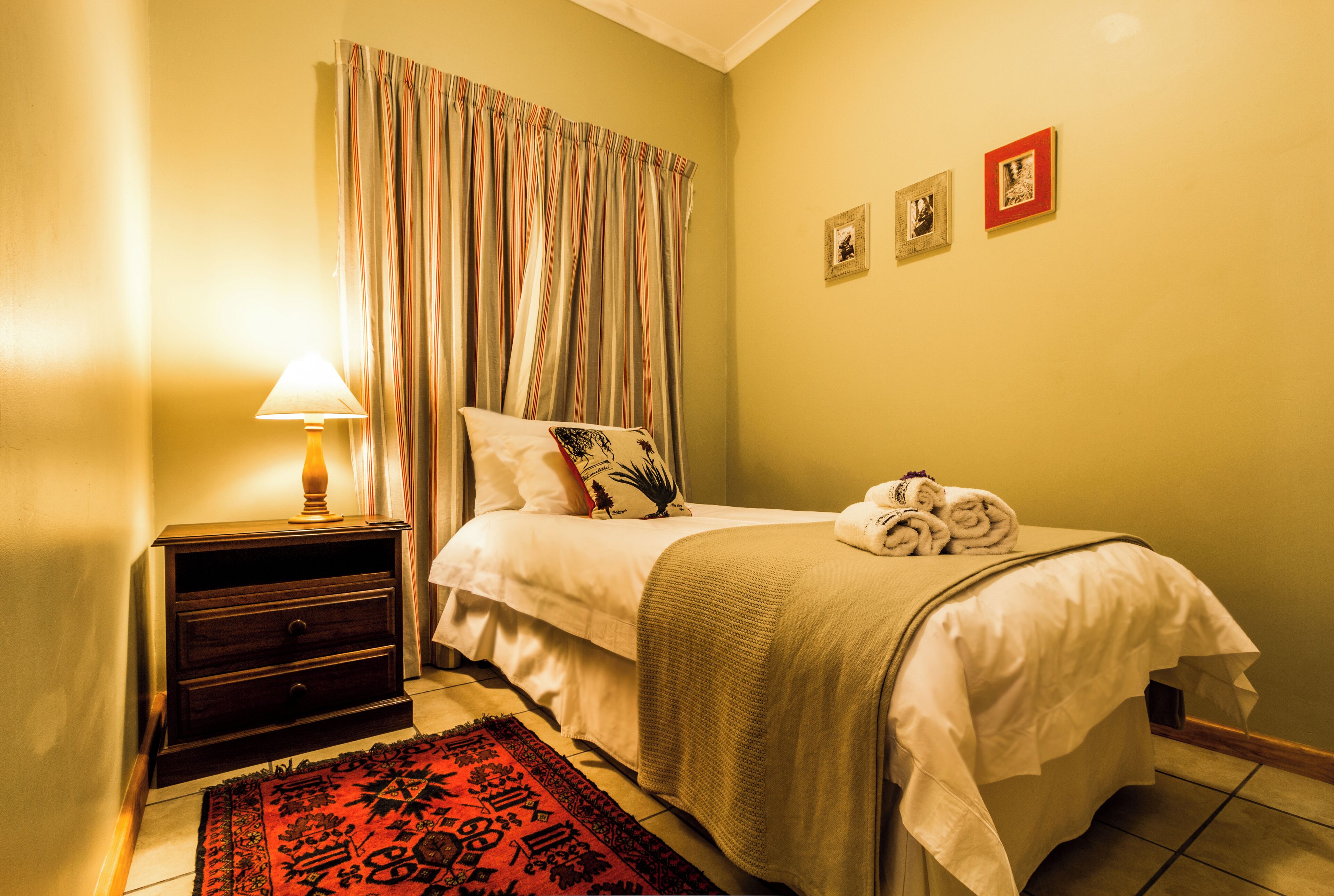 A neatly made bed with a white comforter and a red and black patterned rug, a wooden nightstand with a lamp, and a wall with three framed pictures.