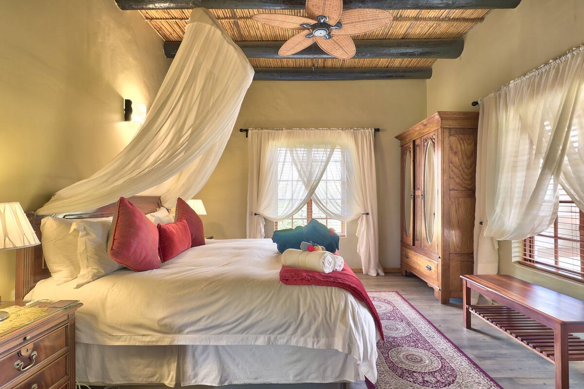 A bedroom with a large bed, a wooden wardrobe, a bench, and a ceiling fan.