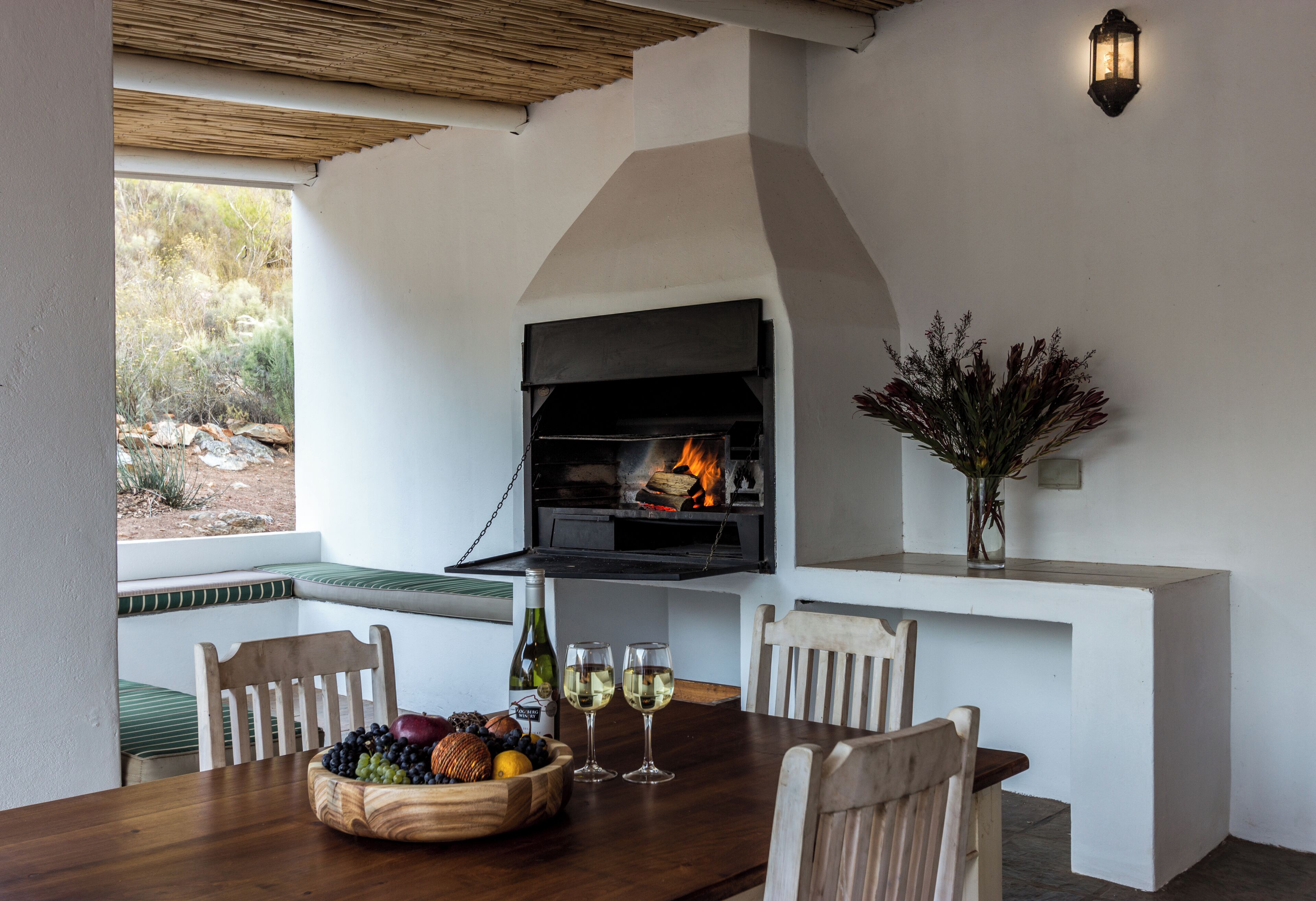 A dining area with a built-in grill, a table set with a fruit bowl and glasses, and a view of the outdoors.
