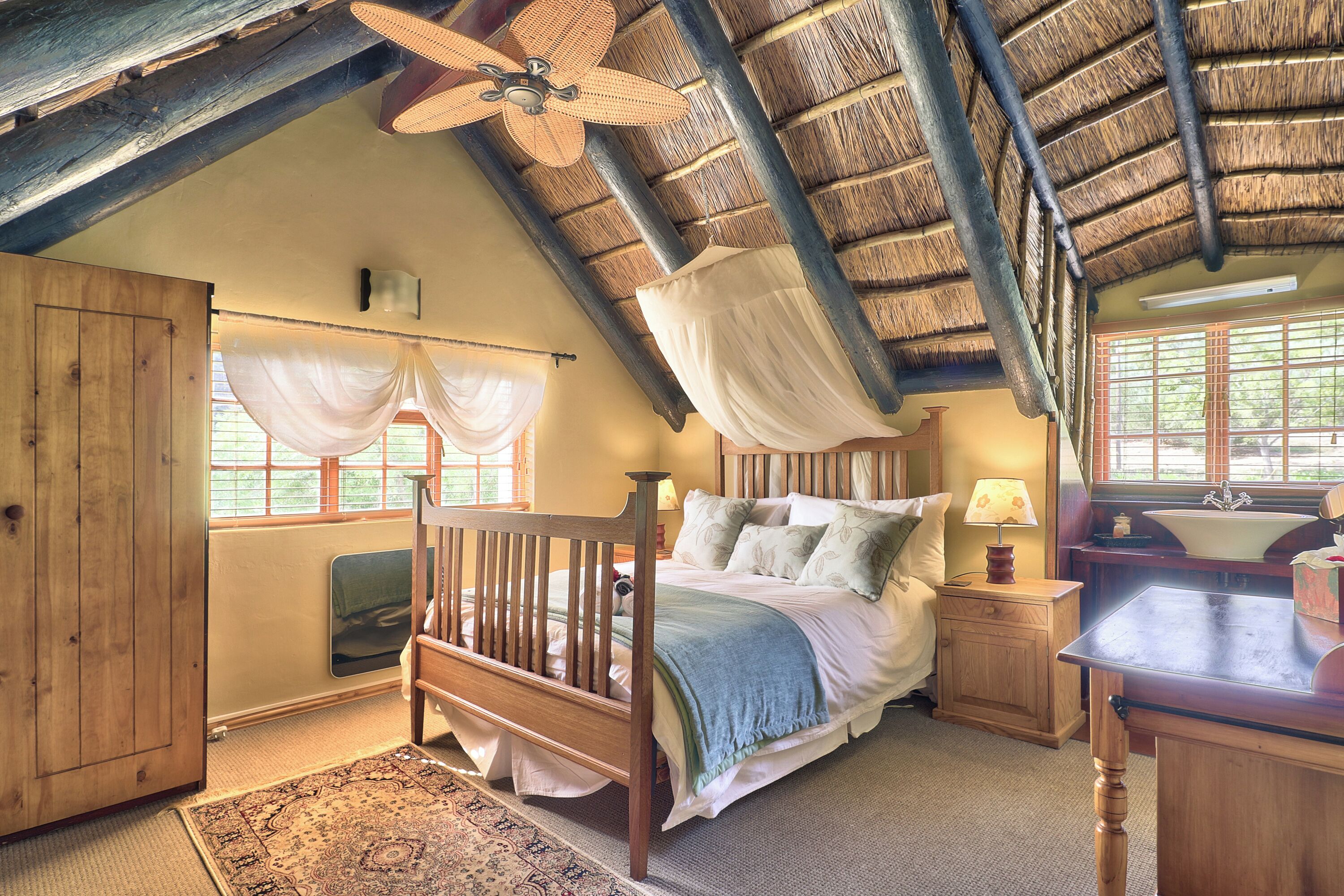 A bedroom with a wooden bed, a nightstand, a window with curtains, a ceiling fan, and a rustic thatched roof.