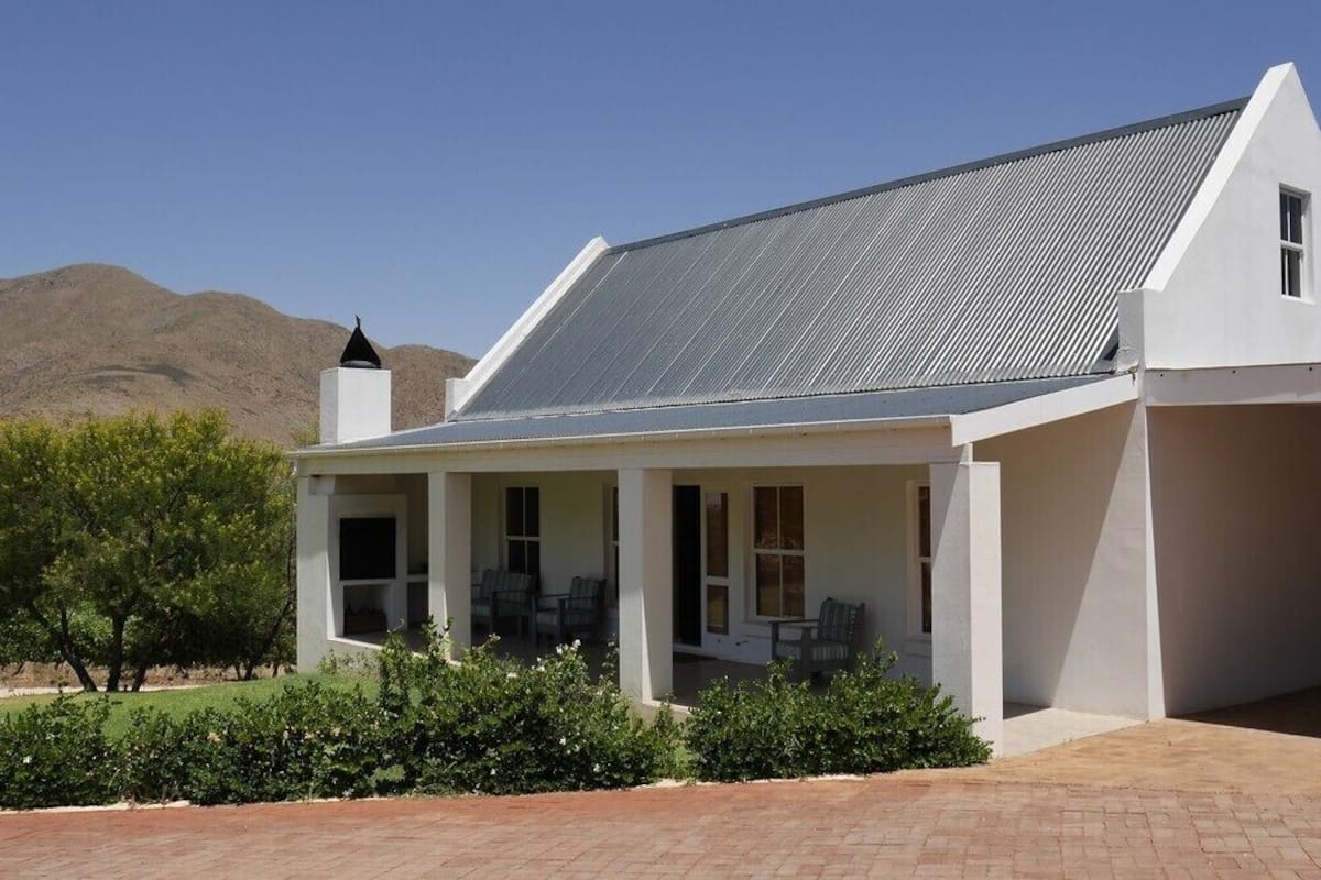 A white building with a sloped roof and a chimney, surrounded by greenery and a clear sky.