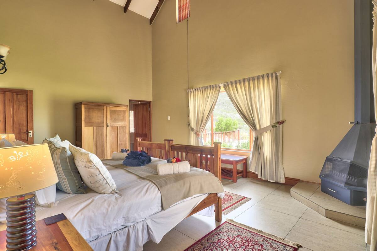 A bedroom with a bed, wooden furniture, a table, a bench, a fireplace, and a view of the outdoors.