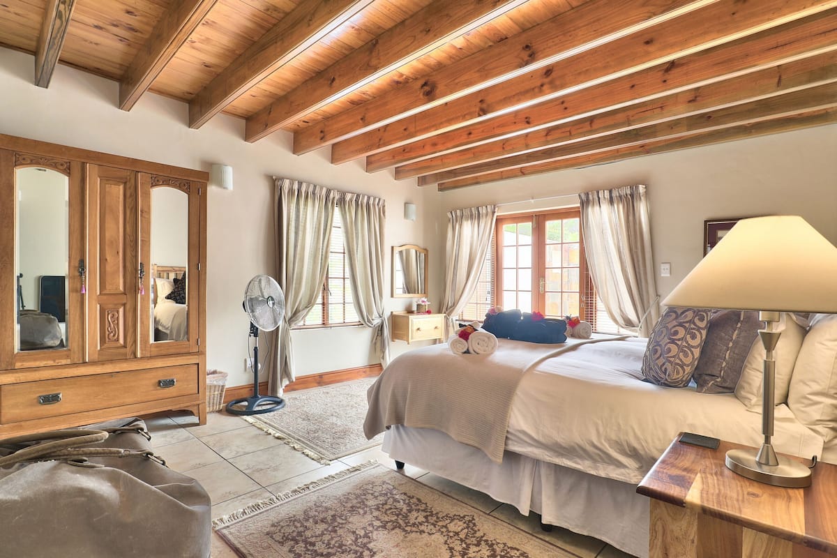 A bedroom with a wooden ceiling, a large bed, a bedside table with a lamp, a dresser, and a mirror.
