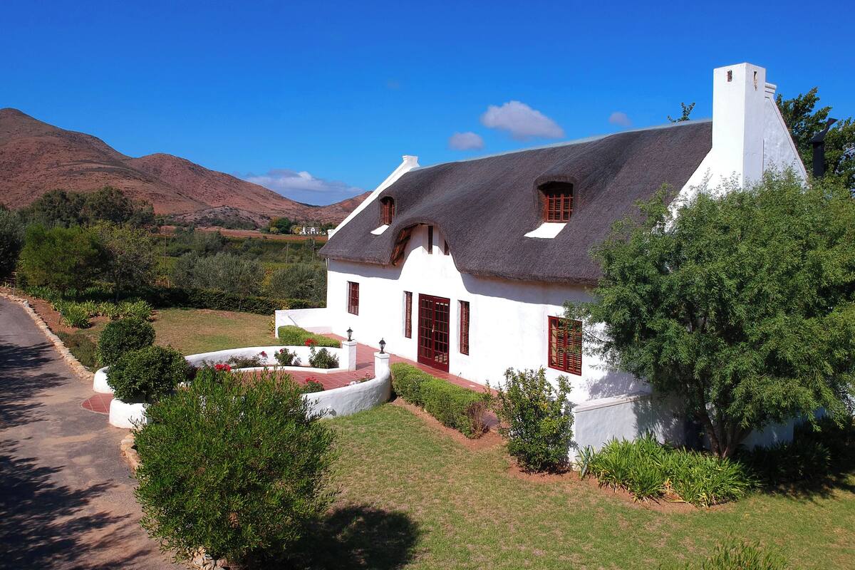 A white house with a thatched roof, surrounded by greenery and hills in the background.
