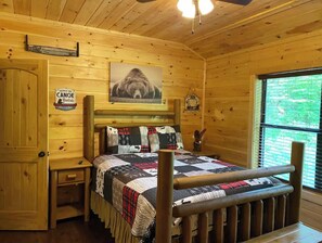 Una habitación acogedora tipo cabaña con interior de madera, una cama con colcha, una mesita de noche, un letrero que anuncia alquiler de canoas y una ventana con vistas a la vegetación.