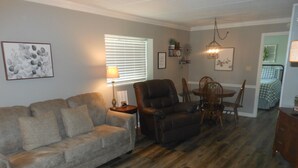 Un salon comprenant un canapé, un fauteuil, une table à manger avec des chaises, une lampe et une fenêtre avec des stores.