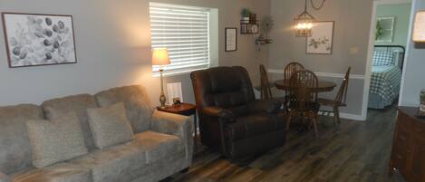 Un salon comprenant un canapé, un fauteuil, une table à manger avec des chaises, une lampe et une fenêtre avec des stores.