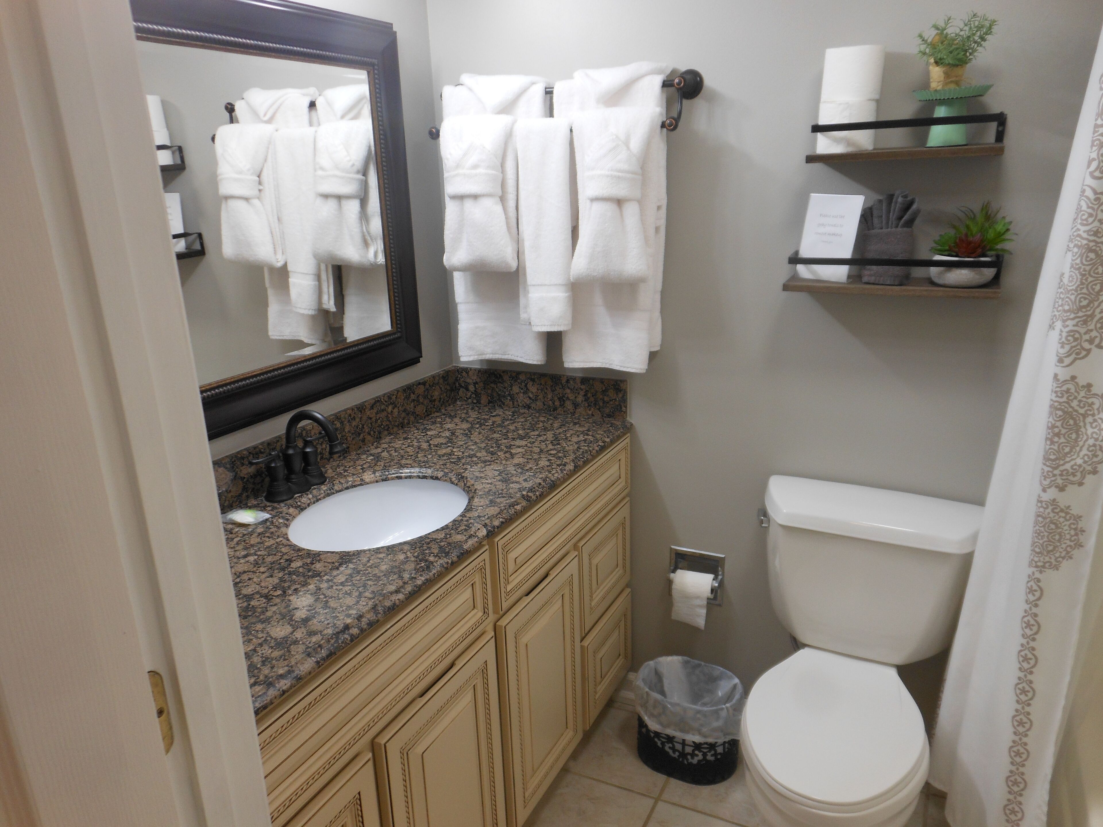 Un baño con lavamanos de encimera de granito, inodoro blanco, un cubo de basura y un espejo.