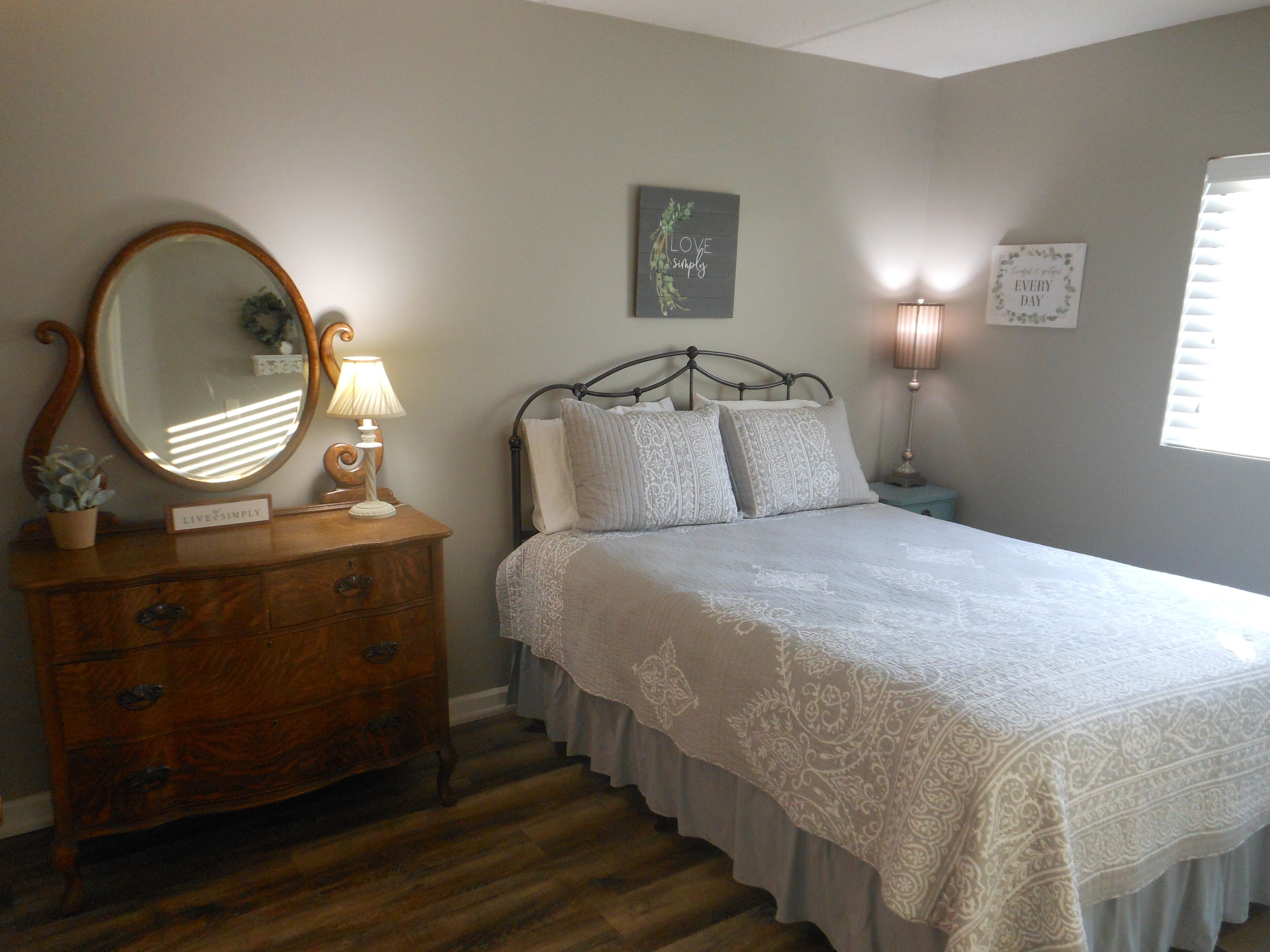 A bedroom with a wooden dresser, a bed with patterned bedding, a mirror, and wall decorations.