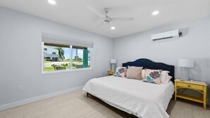 A bedroom with a bed, nightstands, a ceiling fan, an air conditioning unit, and a window with a view of greenery and a building. - Heated Pool Home steps to the beach (Fort Myers Beach)