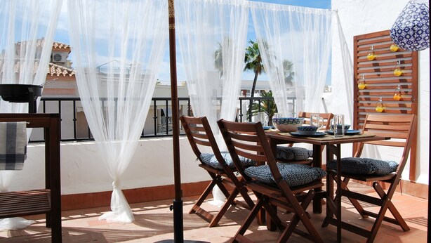 An outdoor patio with a table set for dining, chairs with patterned cushions, a decorative umbrella, and a wall-mounted light fixture. - Ap140 Alcazaba 2D (Nerja)