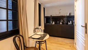 A compact kitchen with a dining area, featuring a table set for two, a chair, and a window with curtains. - Passocean 1 - Suite in the Heart of Honfleur - Kingsize Bed - Airco (Honfleur)