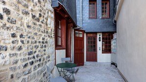 A narrow cobblestone alley with a stone wall on one side and a building with a red door and windows on the other. There is a small table and two chairs outside the building. - Passocean 1 - Suite in the Heart of Honfleur - Kingsize Bed - Airco (Honfleur)