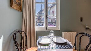 A dining area with a table set for two, a window with a view of buildings, and a radiator on the wall. - Passocean 2 - Elegant Suite in the Heart of Honfleur - Kingsize (Honfleur)