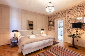 Ein Schlafzimmer mit einem großen Bett, zwei Nachttischlampen, einer Kristallleuchtre, einem gemusterten Wandteppich und einem kleinen Tisch mit Lampe.