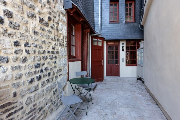 A narrow cobblestone alley with a stone wall on one side and a building with a red door and windows on the other. There is a small table and two chairs outside the building.