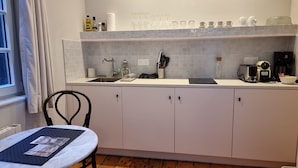 Une cuisine moderne avec un plan de travail blanc, un évier et des placards. On y trouve divers ustensiles de cuisine, comme une machine à café, un grille-pain et un bloc à couteaux. Une chaise et une table sont également visibles.