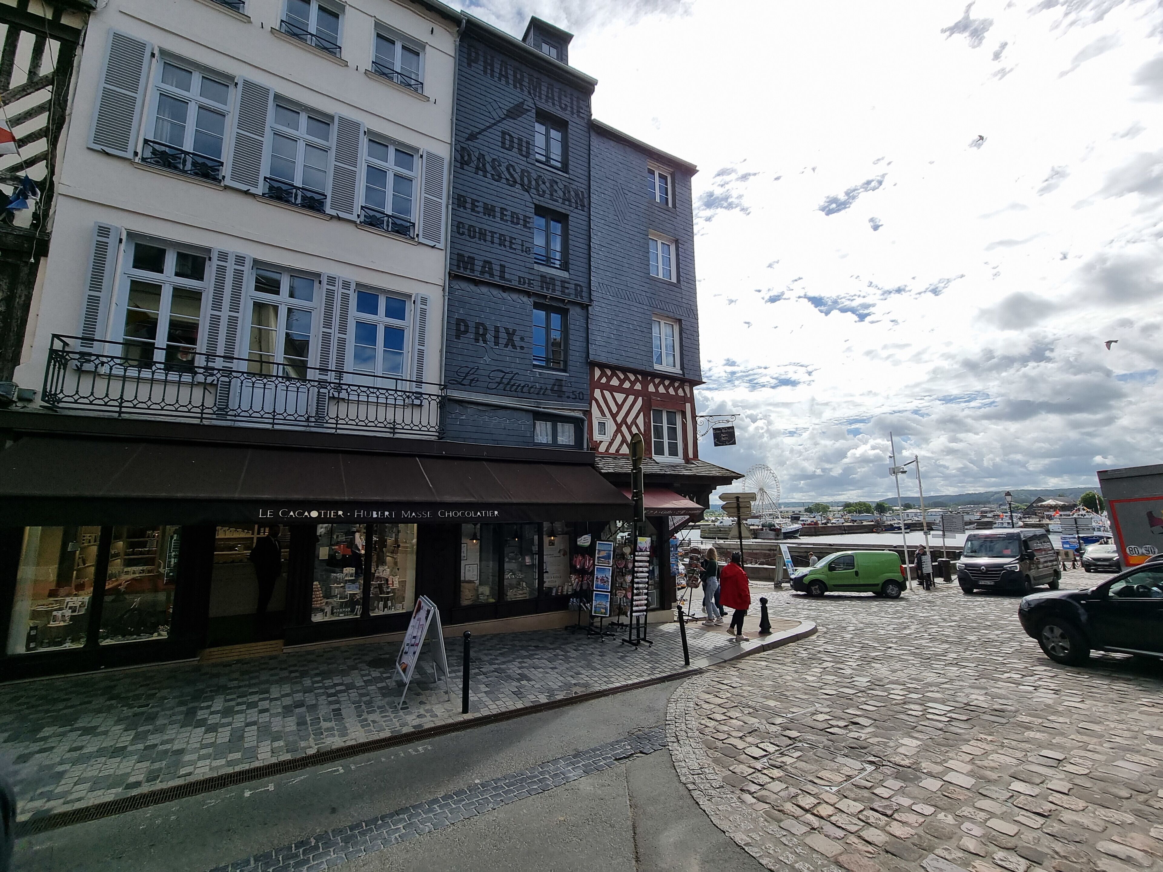 Una calle adoquinada con edificios, un cartel y una noria al fondo.