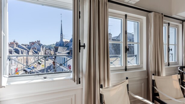 Ein Zimmer mit Blick auf eine Stadtlandschaft, darunter ein Kirchturm, durch große Fenster mit Vorhängen. Zwei Stoffstühle stehen im Raum.