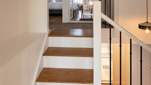 Eine Treppe mit Holzstufen und weißen Geländern, die zu einem Dachzimmer mit Bett und Ausblick nach draußen führt.