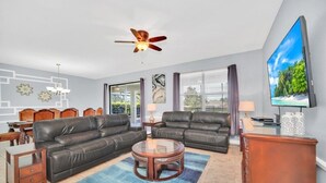 Ein Wohnzimmer mit zwei schwarzen Ledersofas, einem Glastisch, einem Deckenventilator, einem Fernseher und einem Essbereich mit Tisch und Stühlen.