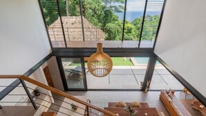 Un intérieur moderne avec un escalier menant à un balcon d’où l’on a vue sur une piscine et une végétation luxuriante.