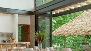 Un espace repas moderne avec une table et des chaises en bois, une grande fenêtre donnant sur un paysage verdoyant et une structure extérieure dotée d’un toit de chaume.