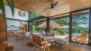 Un espace de vie moderne comprenant un canapé, une table à manger et des chaises, une cuisine en arrière-plan, et une vue sur une piscine et des arbres, le tout visible à travers de grandes fenêtres.