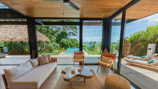 Un salon moderne avec un canapé, une table basse et des fauteuils, de grandes fenêtres offrant une vue sur une piscine et un espace verdoyant.