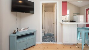 A compact living space with a flat-screen TV mounted on the wall, a light blue cabinet, a small kitchen area with a sink and a refrigerator, and a shower with a curtain.