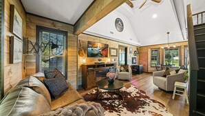A spacious living room with a large sofa, a coffee table, and a TV mounted on the wall. There is a dining area with a table and chairs visible in the background. - Creekside Cabin Smoky Mtn Views Arcade HotTub Pet (Sevierville)