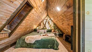 A cozy attic bedroom with a sloped ceiling, a bed with a green bedspread, a small table with a lamp, and a decorative plant arrangement. - Cozy Honeymoon Cabin w/ Mtn Views-Hot Tub-Fire Pit (Sevierville)