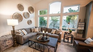A living room with a sectional sofa, a coffee table, and a lamp. There are decorative plates on the wall and a window with a view of trees. - 4br, 3ba, pool near TPC in Scottsdale (Scottsdale)