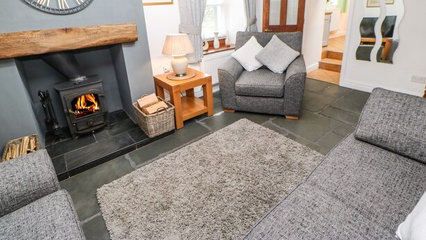 A cozy living room with a fireplace, a grey sofa, a wooden side table, and a large wall clock. - Pine Cottage (Appleby-in-Westmorland)