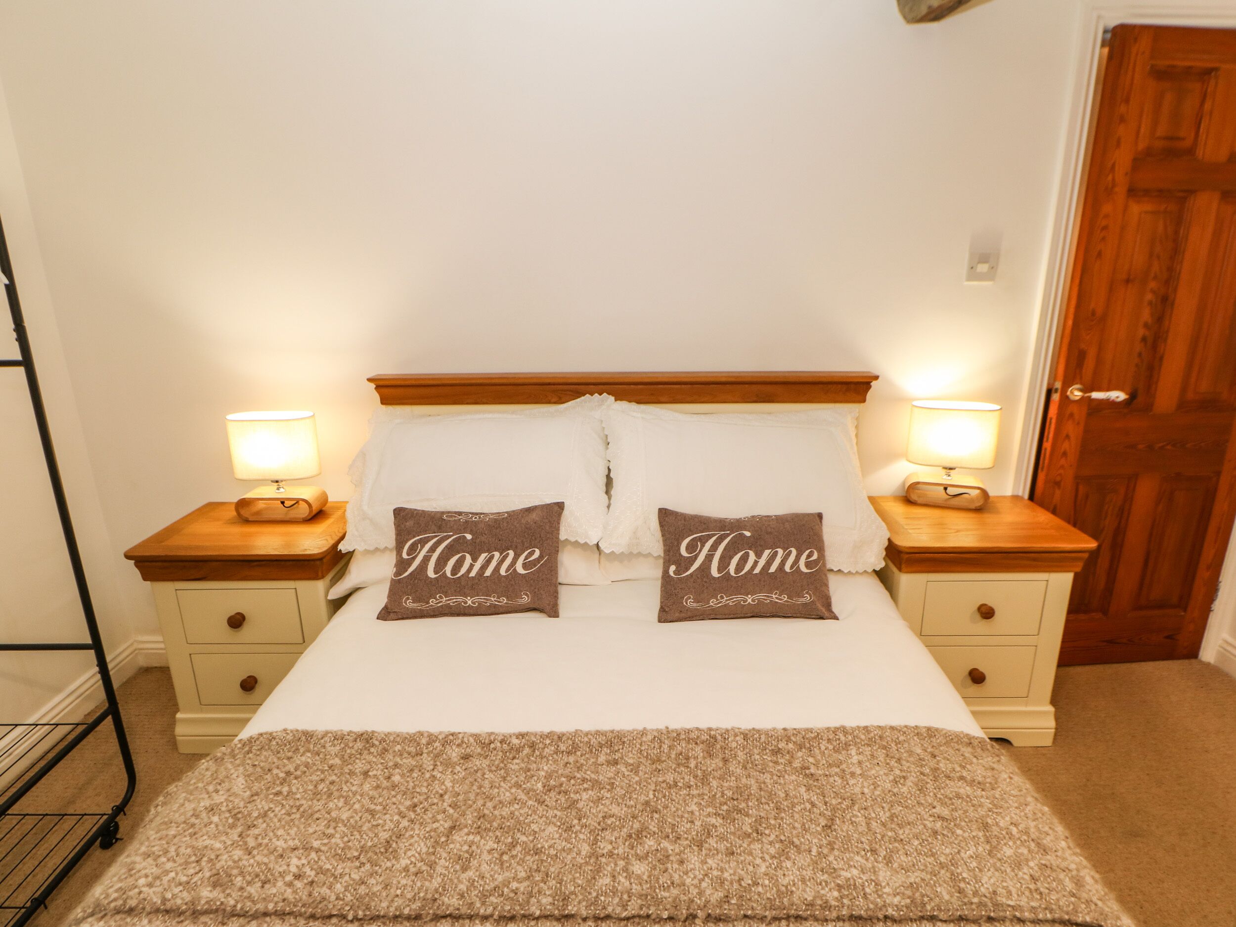 A bedroom with a bed, two bedside tables, two lamps, two pillows with the word 'Home' on them, a wooden headboard, and a door in the background.