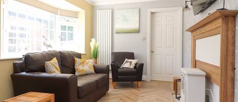 A living room with a brown leather sofa, a wooden coffee table, a black armchair, and a white radiator. There is a television mounted on the wall and a small white cabinet.