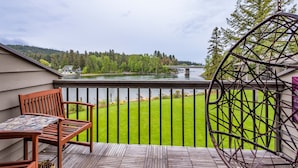 A wooden deck with a chair and table, overlooking a green lawn and a lake with a bridge in the distance. - Waterfront studio w/private balcony & stunning views - Marina Cay Vista Aqua Studio (Bigfork)