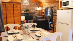 A cozy cabin interior with a dining table set for four, a fireplace, wooden walls, and a white refrigerator.