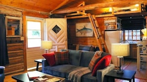 A cozy cabin interior with a wooden ceiling, a sofa with plaid cushions, a dining table, and a ladder leading to a loft area.