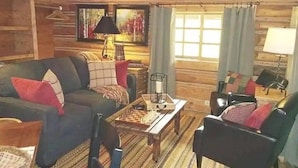 Een gezellig interieur van een houten blokhut met een bank, fauteuils, een salontafel en een raam met gordijnen.